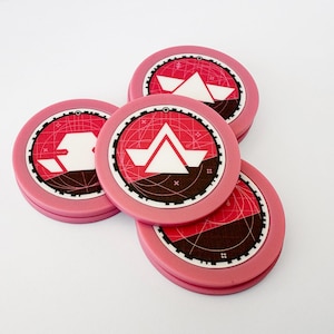 May include: Four pink, circular game tokens with a red and black design. Each token features a white geometric shape in the centre, surrounded by a detailed pattern. The tokens are stacked and slightly overlapping on a white surface.