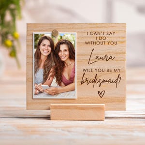 Könnte beinhalten: Ein Holzfotorahmen mit einem Foto von zwei Frauen. Der Rahmen trägt den Text "I can't say I do without you Laura, will you be my bridesmaid?" mit einem Herzsymbol. Er wird von einem kleinen Holzständer getragen.