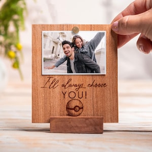 Peut inclure: Cadre photo en bois avec une photo d'un couple. Le cadre porte l'inscription "I'll always choose YOU !" et un dessin de balle. Le cadre est tenu par une main.