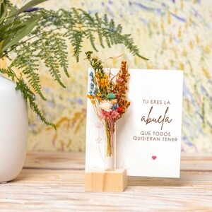 May include: A white card with the text "Tu eres la abuela que todos quisieran tener" and a small heart. A glass tube with dried flowers is in front of the card, which is on a wooden stand.