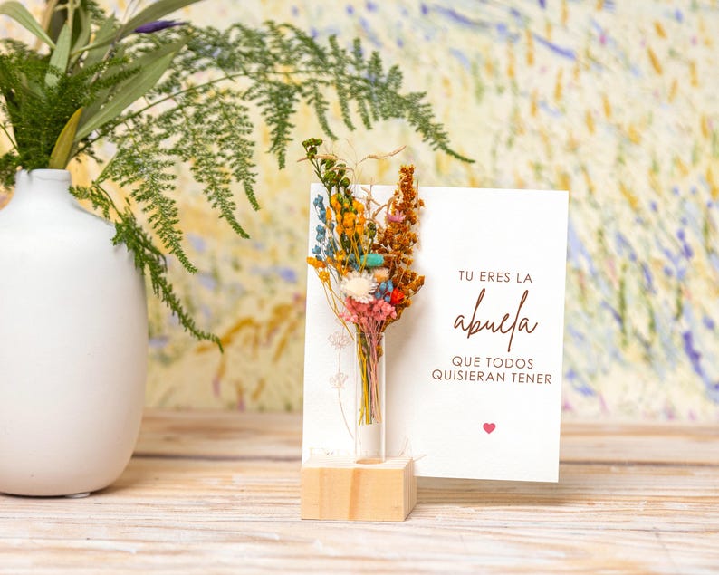 May include: A white card with the text "Tu eres la abuela que todos quisieran tener" and a small heart. A glass tube with dried flowers is in front of the card, which is on a wooden stand.