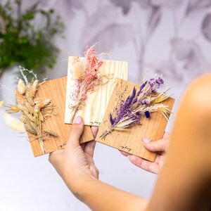 May include: Three wooden cards with dried flower arrangements. The cards are light brown and have the word "everyday" written on them. The flowers are a mix of beige, purple, and pink.
