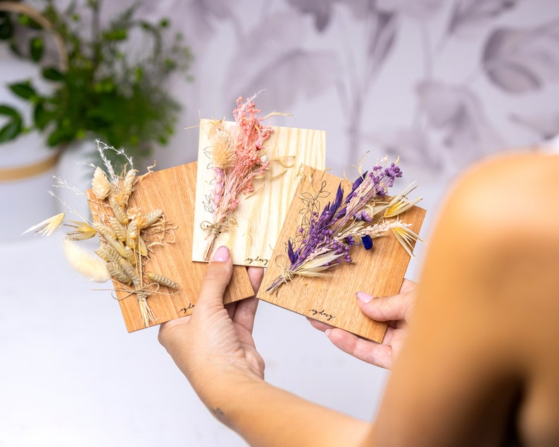 May include: Three wooden cards with dried flower arrangements. The cards are light brown and have the word "everyday" written on them. The flowers are a mix of beige, purple, and pink.