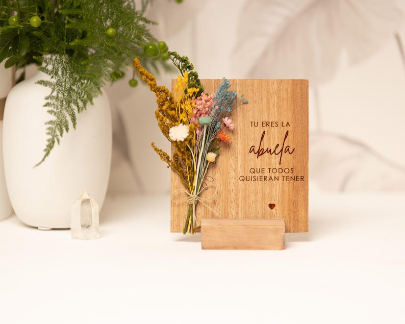 May include: A wooden sign with the text "Tu eres la abuela que todos quisieran tener" (You are the grandmother everyone would want to have) in red lettering. The sign is decorated with dried flowers and a small red heart.