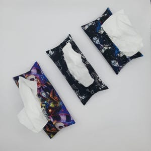 May include: Three rectangular tissue holders with white tissues peeking out. The holders feature colorful, patterned fabric with images of characters. One holder has a blue and purple design, while the others are black with character illustrations.