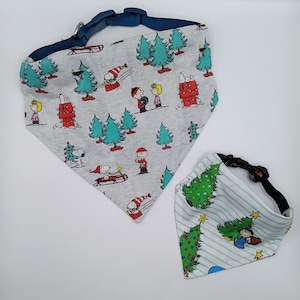 May include: Two triangular pet bandanas with adjustable plastic buckles. One is light gray with a Peanuts holiday print featuring Snoopy, Charlie Brown, and Christmas trees. The other is light blue with white stripes and a Christmas tree print.