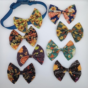 May include: A collection of handmade dog bow ties in various autumn-themed patterns. The bow ties feature sunflowers, pumpkins, and fall leaves in shades of orange, yellow, green, and brown. A blue dog collar is also included in the image.