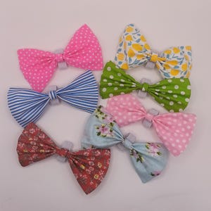 May include: A collection of seven fabric hair bows featuring various patterns including pink with white polka dots, blue and white stripes, yellow lemons, green with white polka dots, light pink with white dots, and two different floral prints.