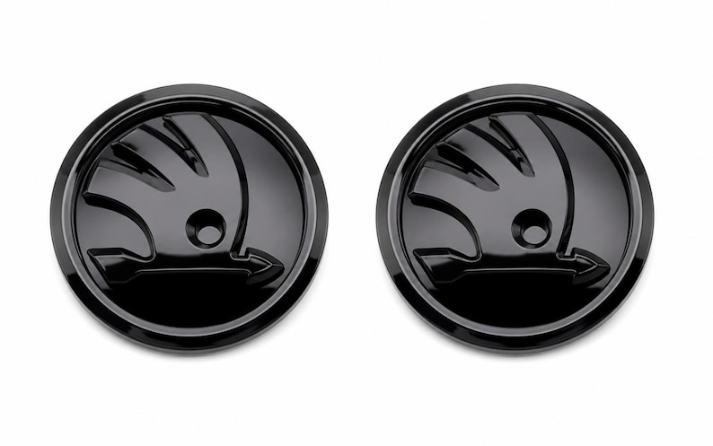 May include: Two glossy black car emblems with the Skoda logo, each in a circular shape. The logo features a stylized winged arrow within a circle. The emblems are set against a white background.