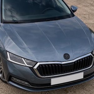 May include: A close-up of a gray-blue car's front end. The car has a black grille, a white license plate, and a black emblem. The headlights are bright white. The car is parked on a gravel surface.