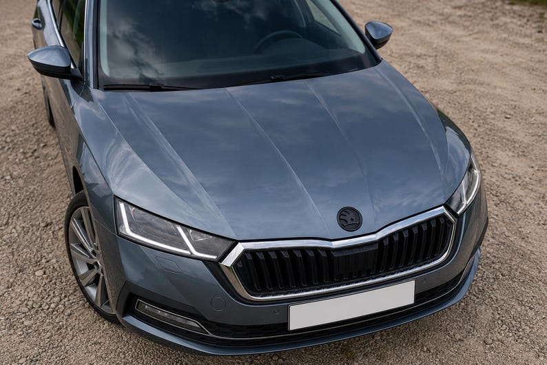 May include: A close-up of a gray-blue car's front end. The car has a black grille, a white license plate, and a black emblem. The headlights are bright white. The car is parked on a gravel surface.
