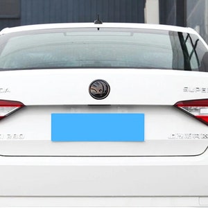 Może przedstawiać: Widok z tyłu białego sedana Skoda Superb. Samoch&oacute;d ma czerwone tylne światła, czarny emblemat i widoczny napis "SUPERB". Obszar tablicy rejestracyjnej jest zakryty niebieskim prostokątem.