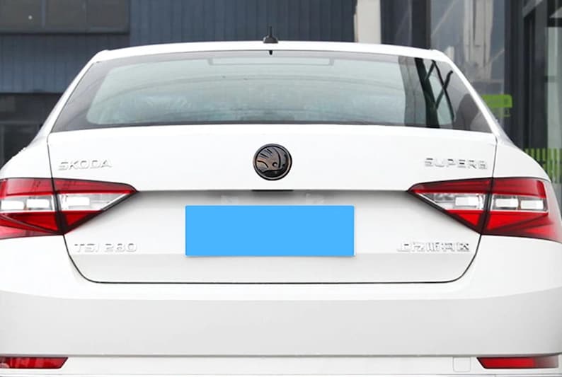 Może przedstawiać: Widok z tyłu białego sedana Skoda Superb. Samoch&oacute;d ma czerwone tylne światła, czarny emblemat i widoczny napis "SUPERB". Obszar tablicy rejestracyjnej jest zakryty niebieskim prostokątem.