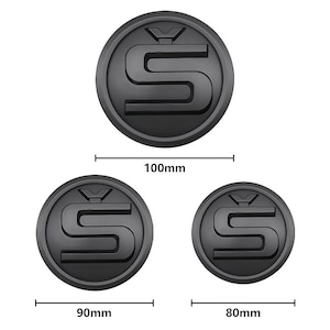 Może przedstawiać: Trzy czarne, okrągłe emblematy ze stylizowanym wzorem "S". Największy emblemat ma średnicę 100 mm, średni 90 mm, a najmniejszy 80 mm. Emblematy mają matowe wykończenie.