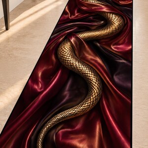 Könnte beinhalten: Ein rechteckiger Läufer mit einem detaillierten goldenen Schlangenmotiv, das sich über einen Hintergrund aus tiefrotem und dunkelviolettem drapiertem Stoff windet. Das Design erzeugt einen kontrastreichen Effekt mit metallischen Schuppen.