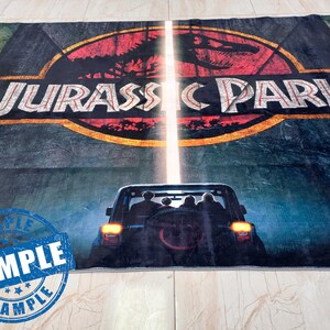 May include: A dark blue rug with the Jurassic Park logo and a silhouette of a jeep driving through a jungle. The logo is in a yellow circle with a red dinosaur inside. The rug has a sample label in the bottom left corner.