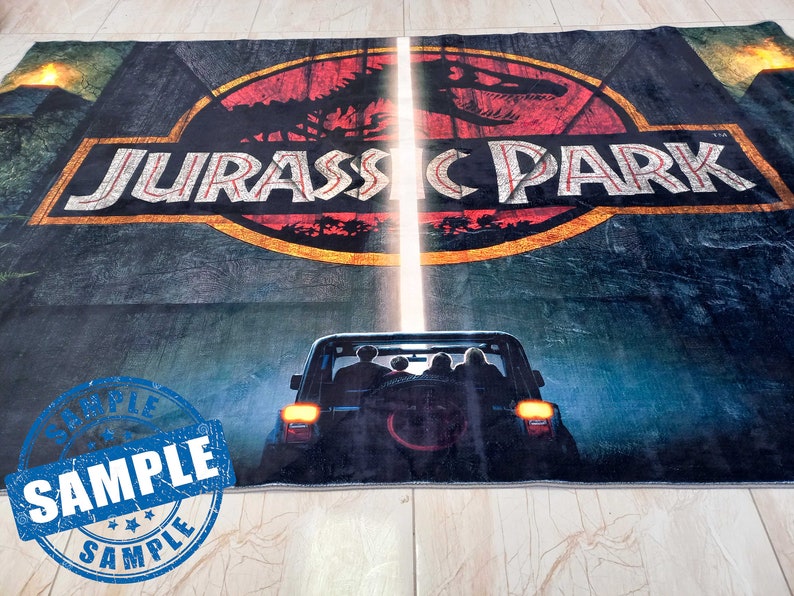 May include: A dark blue rug with the Jurassic Park logo and a silhouette of a jeep driving through a jungle. The logo is in a yellow circle with a red dinosaur inside. The rug has a sample label in the bottom left corner.
