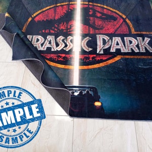 May include: A Jurassic Park themed rug with a dark blue background, a red and yellow logo with a dinosaur silhouette, and the text "Jurassic Park" in white. The rug is partially rolled up, revealing a black backing.