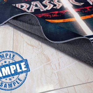 May include: A black and blue rug with a faded red, yellow, and white graphic of the word "RASSICA" on it. The rug is rolled up and the underside is visible, showing a black fabric with a dotted pattern. A blue and white "SAMPLE" stamp is in the foreground.