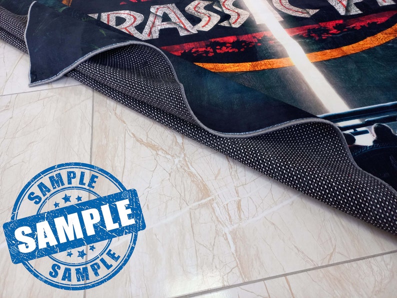 May include: A black and blue rug with a faded red, yellow, and white graphic of the word "RASSICA" on it. The rug is rolled up and the underside is visible, showing a black fabric with a dotted pattern. A blue and white "SAMPLE" stamp is in the foreground.