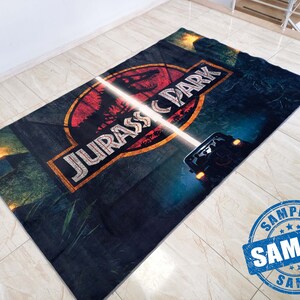 May include: A dark blue rug with a red, yellow, and white Jurassic Park logo. The logo features a dinosaur and the text "Jurassic Park". A white light shines across the rug, illuminating a jeep in the background.