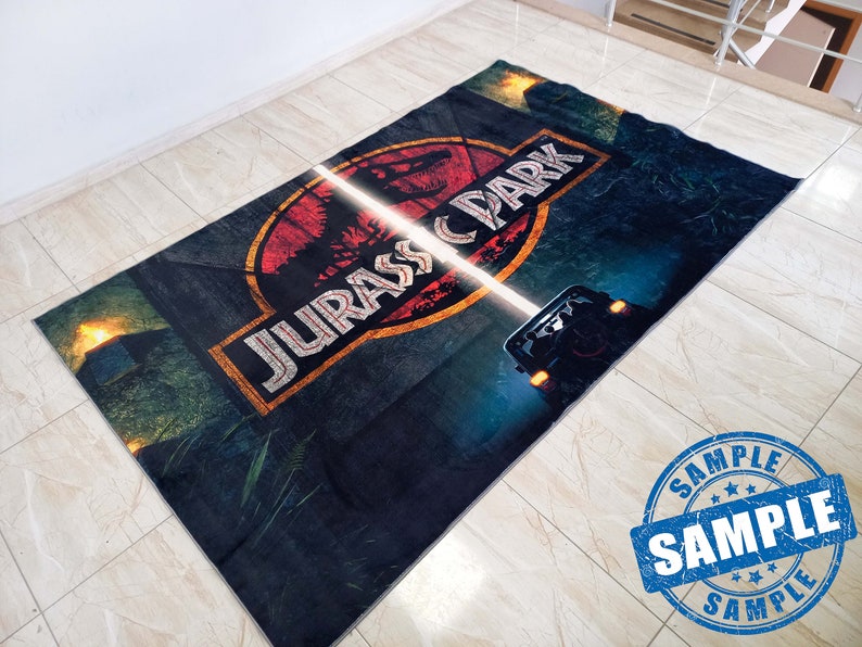 May include: A dark blue rug with a red, yellow, and white Jurassic Park logo. The logo features a dinosaur and the text "Jurassic Park". A white light shines across the rug, illuminating a jeep in the background.