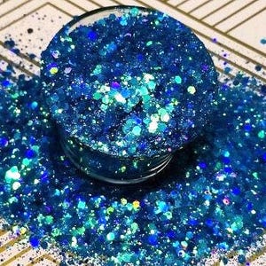 May include: A close-up of a jar filled with blue and teal iridescent glitter. The glitter is scattered around the jar, creating a sparkling effect.