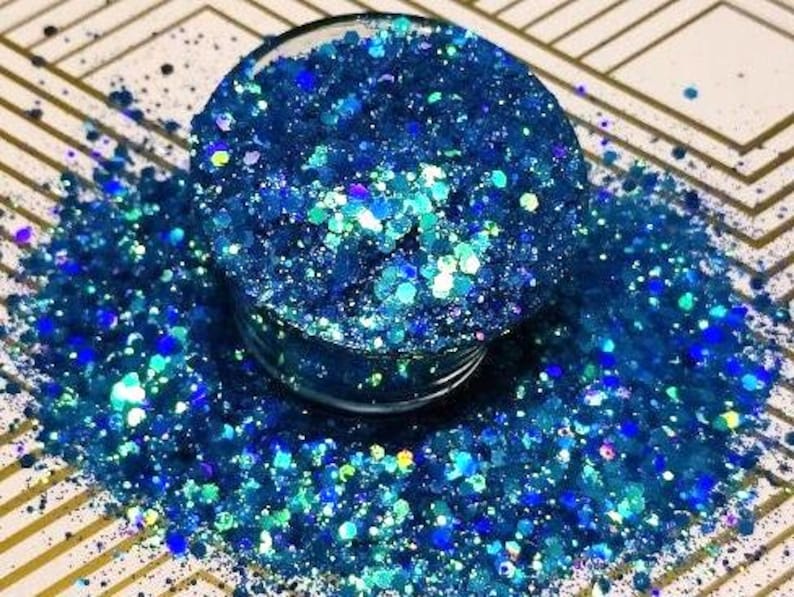 May include: A close-up of a jar filled with blue and teal iridescent glitter. The glitter is scattered around the jar, creating a sparkling effect.