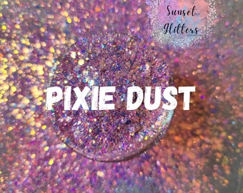 May include: A close-up of a small jar of iridescent glitter labeled "Pixie Dust". The glitter is a mix of pink, purple, blue, and gold. The jar is sitting on a bed of iridescent glitter.