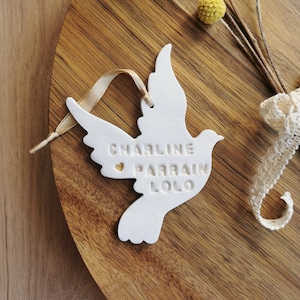 May include: White ceramic dove ornament with the text "CHARLINE PARRAIN LOLO" and a small gold heart. The ornament is hanging from a gold ribbon and is on a wooden surface.