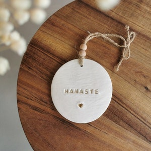 May include: White ceramic ornament with the word "Namaste" and a small heart in gold. The ornament is hanging from a brown twine string with wooden beads.