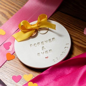 May include: A white ceramic ring dish with the words "Forever & Ever" in gold lettering. The dish is decorated with a yellow bow and two gold rings. The dish is surrounded by pink and orange confetti hearts on a wooden surface.