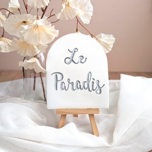 Op de afbeelding: Wit boogvormig bord op een klein houten ezel, met de woorden "Le Paradis" in een cursief lettertype. Het bord staat tegen een achtergrond van witte stof en crèmekleurige bloemen.