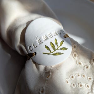 Puede incluir: Servilletero de cerámica blanca con el nombre "CLEMENCE" en relieve y un diseño de rama de olivo pintada. El anillo está asegurado con un cordón marrón y descansa sobre una servilleta blanca con detalles calados.