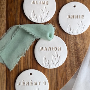 Puede incluir: Etiquetas de cerámica blanca con nombres grabados y diseños de hojas. Se ven los nombres "Nicolas" y "Aline". Una cinta verde salvia y una superficie de madera completan la escena. Perfectas para eventos.