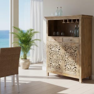 May include: A light-colored wooden cabinet with carved floral designs on the doors. The cabinet has a top section for glasses and bottles, and two drawers. A potted plant and a chair are in the foreground.