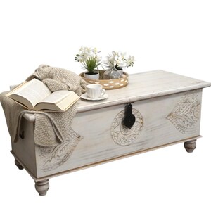 May include: White wooden storage chest with carved details and a black metal latch. The chest is sitting on a white surface and has a book, a cup, and a tray of flowers on top.