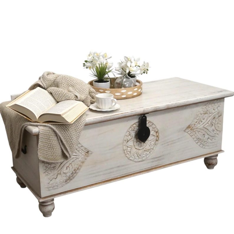 May include: White wooden storage chest with carved details and a black metal latch. The chest is sitting on a white surface and has a book, a cup, and a tray of flowers on top.