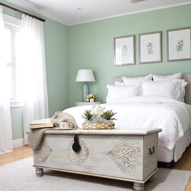 May include: A white wooden storage trunk with intricate carvings sits in front of a bed with white bedding and a light green wall. The trunk has a black metal latch and two handles. There are plants in a wooden tray on top of the trunk.