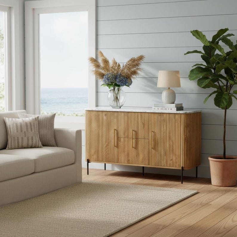 K&ouml;nnte beinhalten: Ein helles Holzschrank mit einer wei&szlig;en Marmorplatte, drei T&uuml;ren und schwarzen Beinen. Der Schrank befindet sich in einem Wohnzimmer mit einem beigen Teppich, einem Sofa und einem gro&szlig;en Fenster mit Blick auf das Meer. Eine Lampe und eine Blumenvase stehen oben.