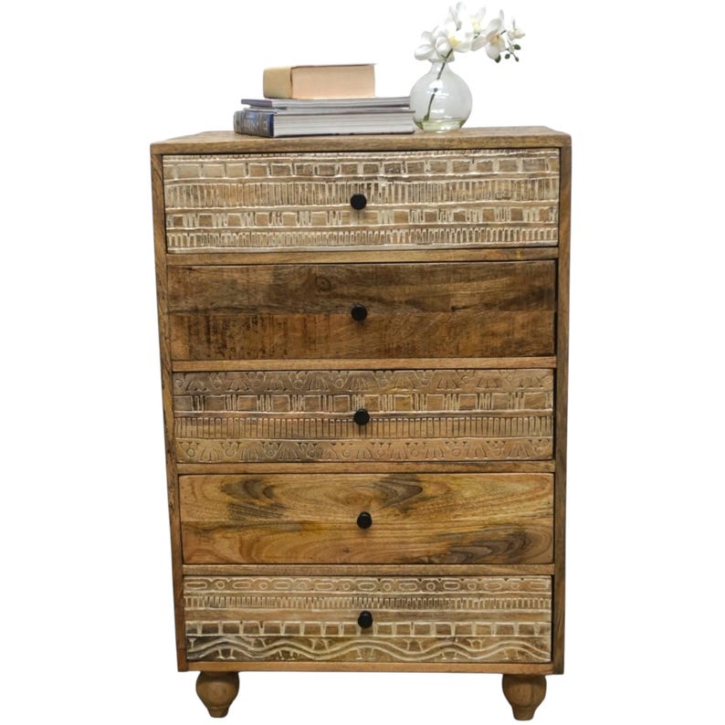 May include: A wooden chest of drawers with five drawers. The top, third, and bottom drawers have a carved, light-coloured design. The other drawers are a darker wood. The chest has small, round feet and black knobs.