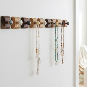May include: A wooden wall-mounted jewelry organizer with eight square blocks in varying shades of brown. Each block has a decorative knob, and several necklaces of different styles hang from the knobs. The wall is white.