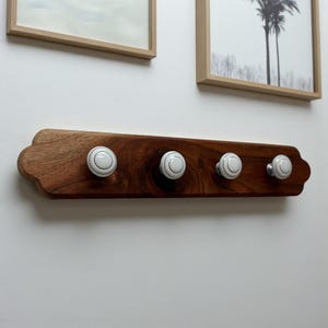 May include: A wooden coat rack with four white ceramic knobs. The rack is made of dark brown wood and has a decorative edge. The knobs are round with a white and gray pattern. The rack is mounted on a white wall.