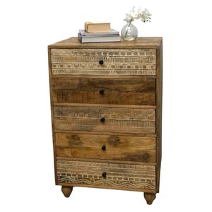 May include: A wooden chest of drawers with five drawers. The top and bottom drawers have a carved, patterned design. The other drawers are plain. The chest is topped with books and a vase of white flowers. The chest is made of wood and has a natural finish.
