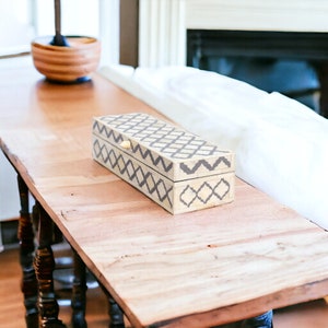 May include: A rectangular decorative box with a white and gray geometric pattern. The box is made of bone and has a brass handle. The box is sitting on a wooden table.
