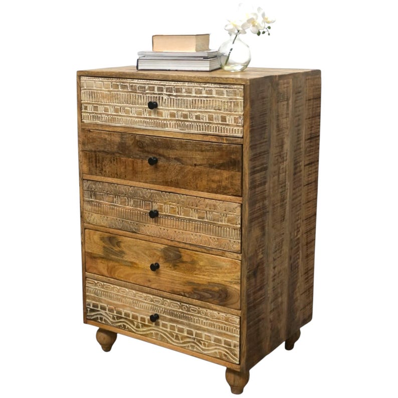 May include: A wooden chest of drawers with five drawers. The top and bottom drawers have a white carved design. The other drawers are a medium brown colour. The chest has black knobs and sits on four small, rounded feet. A vase of white flowers and books are on top.