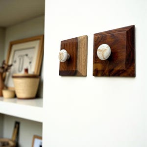 May include: Two wooden wall hooks with a square design. The hooks are made of dark brown wood with a light-colored, patterned knob. The wood has a natural grain, and the knobs are off-white with brown markings.