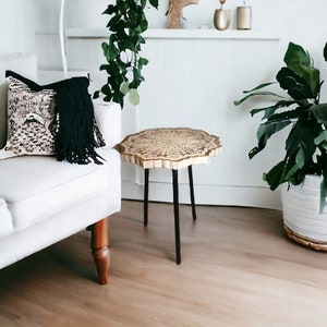 May include: A small, round side table with a carved wooden top and black metal legs. The table has a mandala design on the top and is sitting on a light wood floor.