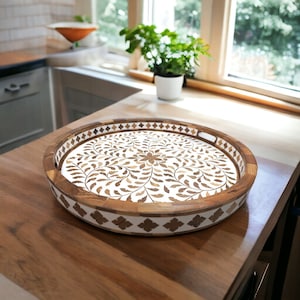 May include: Round wooden serving tray with a white and brown floral design. The tray has a dark brown rim and a white border with a geometric pattern. The tray is on a wooden surface.