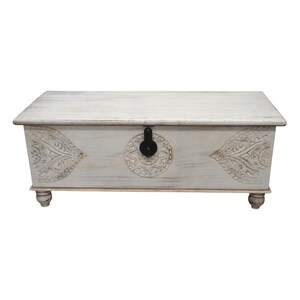 May include: White wooden storage chest with carved floral designs and a black metal latch. The chest has a distressed white finish and sits on four turned legs.
